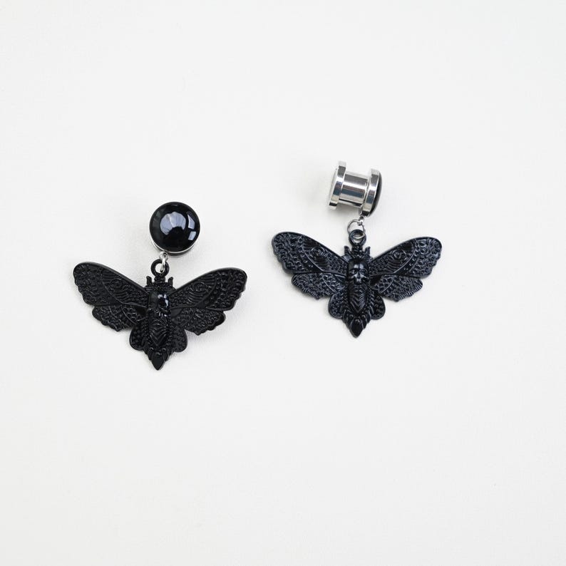 May include: Two black butterfly-shaped ear gauges. One gauge has a black, round end, while the other has a silver, cylindrical end. Both have a detailed butterfly pendant hanging from them. The butterflies have intricate wing patterns.