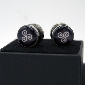 May include: A pair of silver cufflinks with a black background and a white triquetra symbol.