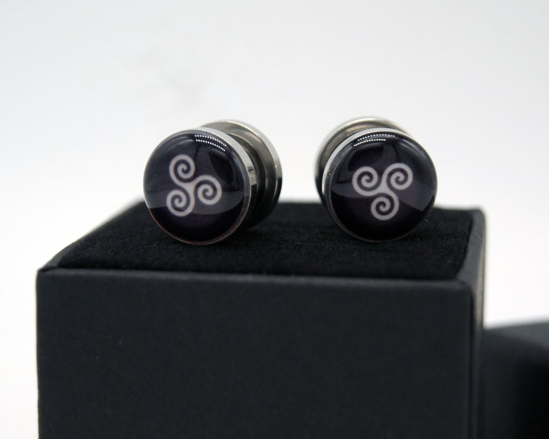 May include: A pair of silver cufflinks with a black background and a white triquetra symbol.