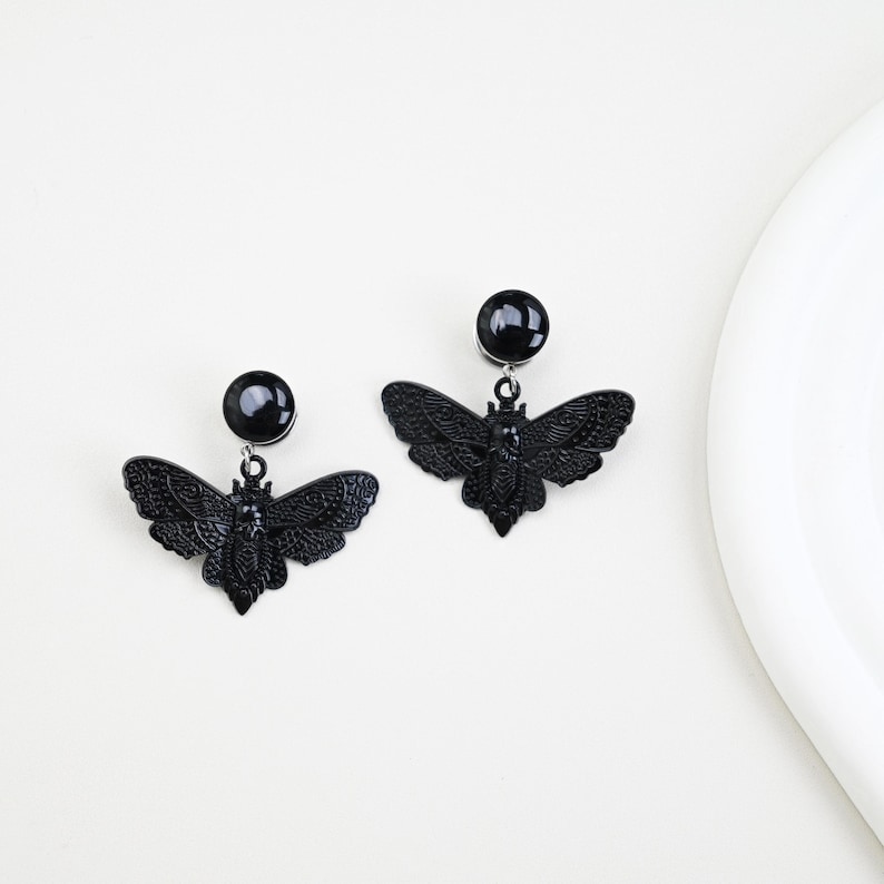 May include: A pair of black earrings featuring a moth design. Each earring has a round stud and a detailed moth pendant. The earrings are set against a white background, highlighting the dark, gothic aesthetic.
