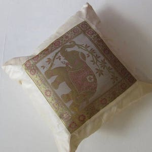 May include: A cream-colored pillow with a gold and brown embroidered elephant design. The elephant is surrounded by floral motifs and the pillow has a satin border.