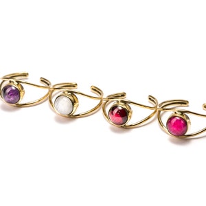 May include: Four gold-toned bracelets with different coloured gemstones. The bracelets are made of thin, curved metal and feature a single round gemstone in the centre. The gemstones are purple, white, red, and pink.