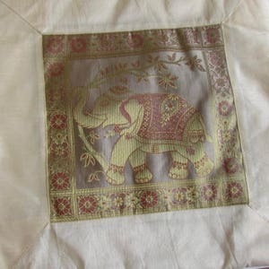 May include: A white throw pillow with a gold and brown embroidered elephant design. The pillow has a cream colored background and a decorative border.