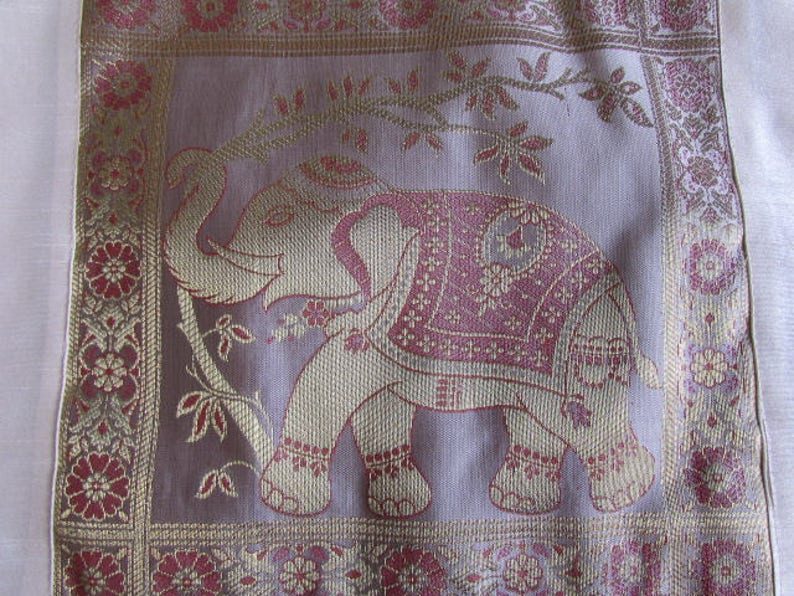 May include: A pink and gold embroidered fabric with an elephant design. The elephant is outlined in gold thread and has pink and gold details. The fabric has a floral border.