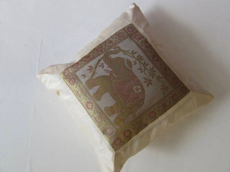 May include: A cream-colored pillow with a gold and brown embroidered elephant design. The elephant is surrounded by floral motifs and the pillow has a satin border.