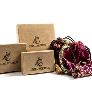 Puede incluir: Tres cajas de cart&oacute;n marr&oacute;n con el logotipo "Gecko Spiritus" y un dise&ntilde;o de gecko negro. Las cajas est&aacute;n apiladas una encima de la otra. Una bolsa de tela con estampado morado y dorado est&aacute; delante de las cajas.