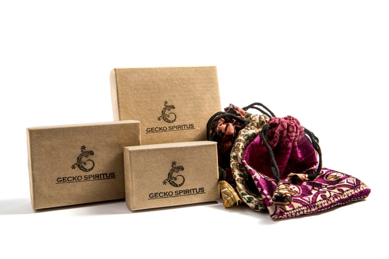 Puede incluir: Tres cajas de cart&oacute;n marr&oacute;n con el logotipo "Gecko Spiritus" y un dise&ntilde;o de gecko negro. Las cajas est&aacute;n apiladas una encima de la otra. Una bolsa de tela con estampado morado y dorado est&aacute; delante de las cajas.