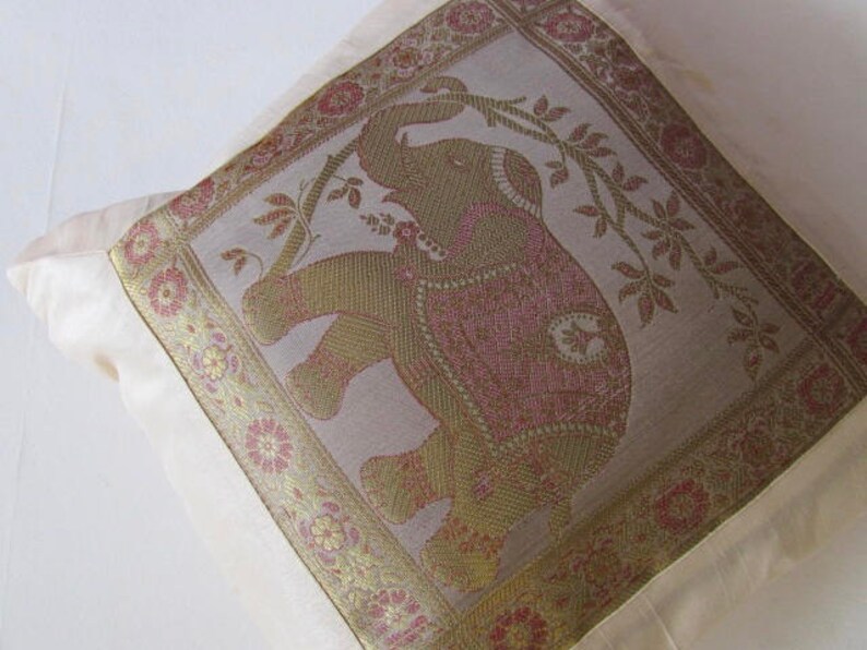 May include: A white throw pillow with a gold and pink embroidered elephant design. The elephant is surrounded by floral patterns and vines.