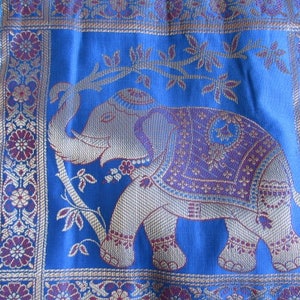 May include: A blue fabric with a gold and purple embroidered elephant design. The elephant is standing on a branch with leaves. The fabric has a border of gold and purple flowers.