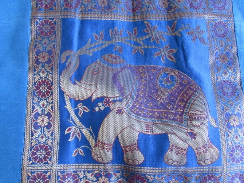 May include: A blue fabric with a gold and purple embroidered elephant design. The elephant is standing on a branch with leaves. The fabric has a border of gold and purple flowers.