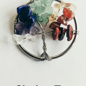 Puede incluir: Un colgante de &aacute;rbol de la vida envuelto en alambre plateado con siete piedras de chakra. Las piedras son transparentes, amatista, azules, verdes, amarillas, naranjas y rojas.