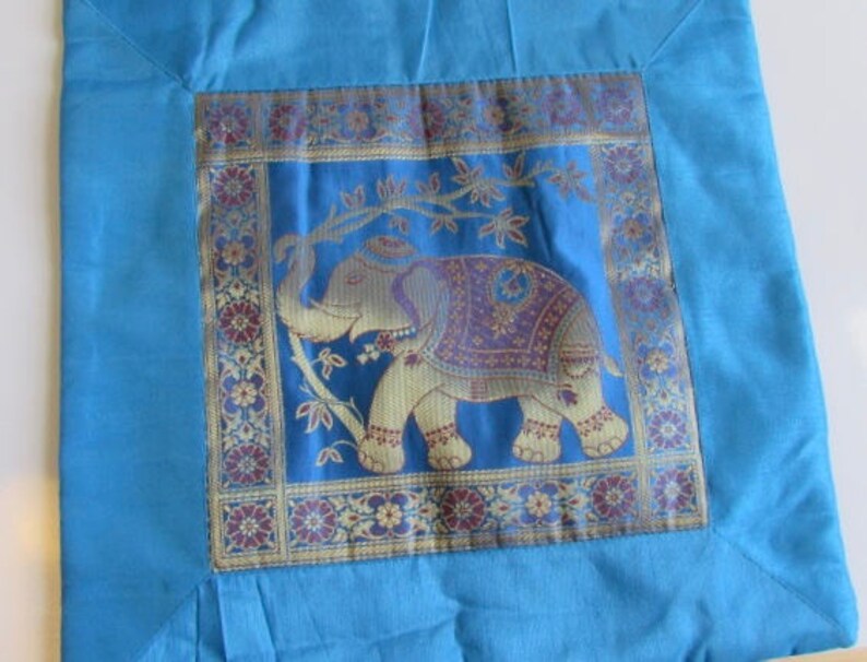 May include: A blue throw pillow with a square embroidered panel featuring a gold and purple elephant on a blue background. The panel is surrounded by a gold and burgundy border with floral designs.