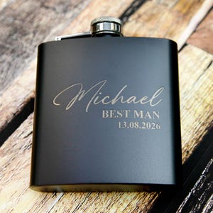 May include: A black hip flask with a silver cap. The flask is engraved with the name "Michael", the words "BEST MAN", and the date "13.08.2026" in a light colour. The flask is rectangular and rests on a wooden surface.