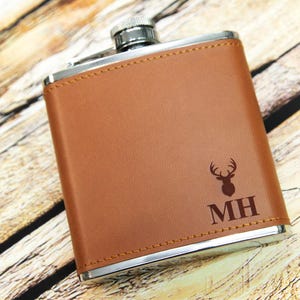 May include: A brown leather-wrapped flask with a silver metal body. The flask has a deer head design and the initials "MH" engraved on the front.