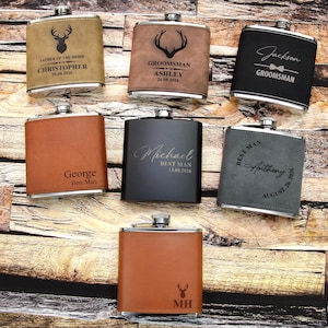 Puede incluir: Una colección de petacas personalizadas en varios colores, incluyendo marrón, negro y gris. Cada petaca presenta texto y diseños personalizados, como una silueta de ciervo o el nombre "Jackson". Las petacas están dispuestas sobre una superficie de madera.