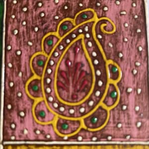 K&ouml;nnte beinhalten: Eine rechteckige dekorative Fliese mit einem zentralen gelben Paisley-Motiv, das mit Reliefmalerei hervorgehoben ist. Der Hintergrund ist in einem strukturierten Altrosa gehalten und mit kleinen wei&szlig;en Punkten sowie gr&uuml;nen Kreisen verziert.