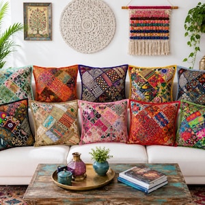 Puede incluir: Una colección de diez cojines cuadrados dispuestos sobre un sofá blanco. Cada cojín presenta un diseño de patchwork único compuesto por diversos tejidos coloridos, bordados intrincados, pequeños espejos y costuras decorativas variadas.