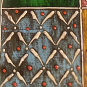 K&ouml;nnte beinhalten: Eine quadratische Keramikfliese mit blauem Hintergrund und einem sich wiederholenden Muster aus erhabenen wei&szlig;en V-f&ouml;rmigen Linien und kleinen orangefarbenen Punkten. Die Fliese ist von dunklen Linien und angrenzenden gr&uuml;nen und gelben Feldern umrandet.