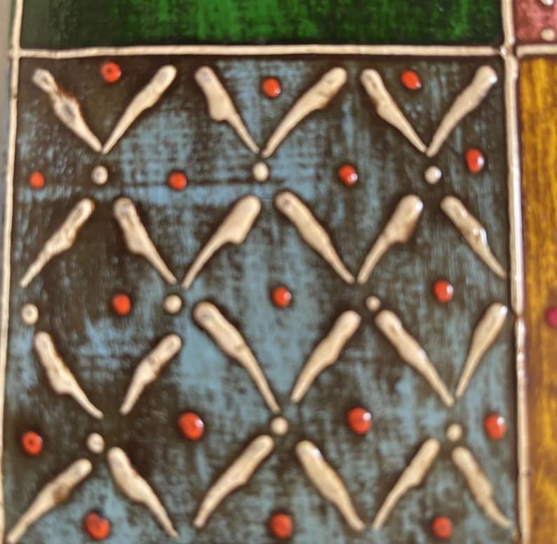 K&ouml;nnte beinhalten: Eine quadratische Keramikfliese mit blauem Hintergrund und einem sich wiederholenden Muster aus erhabenen wei&szlig;en V-f&ouml;rmigen Linien und kleinen orangefarbenen Punkten. Die Fliese ist von dunklen Linien und angrenzenden gr&uuml;nen und gelben Feldern umrandet.