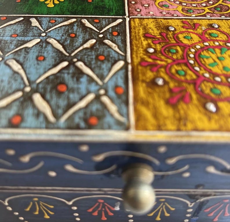K&ouml;nnte beinhalten: Eine Holzschachtel mit einer mehrfarbigen Oberfl&auml;che aus quadratischen Feldern in Blau, Gr&uuml;n, Gelb und Pink, verziert mit wei&szlig;en, orangen und gr&uuml;nen Blumen- und Geometriemustern. Die Vorderseite ist dunkelblau mit wei&szlig;en Linien und einem Knauf.