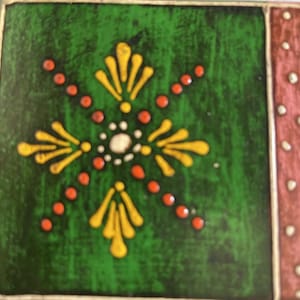 K&ouml;nnte beinhalten: Eine quadratische Keramikfliese mit tiefgr&uuml;nem Hintergrund und einem zentralen gelb-wei&szlig;en Blumenmotiv. Das Design umfasst strahlenf&ouml;rmige orangefarbene Punkte und gelbe Bl&uuml;tenblattformen. Ein vertikaler Rand rechts ist rosa mit wei&szlig;en Punkten.