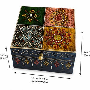 K&ouml;nnte beinhalten: Eine quadratische Holzschatulle mit Klappdeckel, die vier verschiedene bemalte Felder in Gr&uuml;n, Rosa, Blau und Gelb mit komplexen Blumen- und Geometriemustern aufweist. Die dunkelblaue Vorderseite hat einen Messingknopf und misst 15 cm Breite, 15 cm Tiefe und 8 cm H&ouml;he.