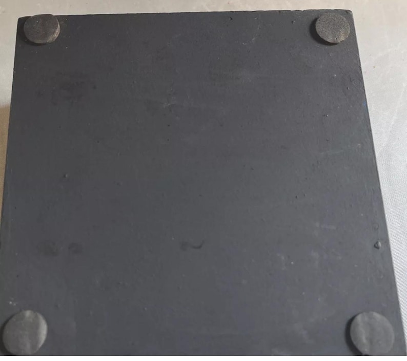 K&ouml;nnte beinhalten: Eine quadratische, mattschwarze Basis mit einer glatten, flachen Oberfl&auml;che. An jeder der vier Ecken auf der Unterseite sind kleine, kreisf&ouml;rmige, dunkelgraue Filz- oder Gummipolster angebracht, die f&uuml;r Stabilit&auml;t sorgen und Kratzer verhindern.