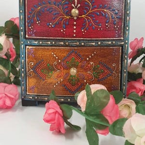 以下が含まれることがあります： 手描きの花柄が施された、2段の木製ボックス。上の引き出しは赤色で、青と金のアクセントがあり、下の引き出しは茶色で、緑と金のアクセントがあります。ボックスは青いフレームと金のノブが付いています。ピンクのバラがボックスを囲んでいます。