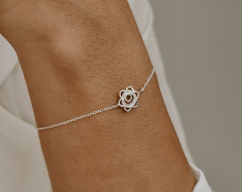 May include: A delicate silver chain bracelet featuring a small, symmetrical, six-petaled floral charm at the center. The metallic charm has a hollow, circular core and pointed petal edges, connected to a thin, polished silver link chain.