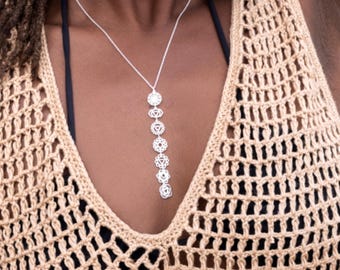 May include: A silver-toned necklace featuring a vertical pendant composed of seven distinct circular and triangular chakra symbols arranged in a linear sequence, suspended from a delicate chain against a beige crocheted garment.