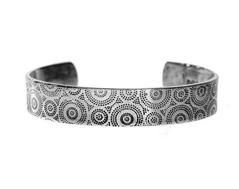 May include: A silver cuff bracelet with a repeating pattern of dotted circles. The open-ended bracelet is made of a shiny metal and features a detailed design across its surface. The bracelet is shown against a white background.