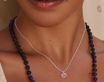 May include: A silver chain necklace featuring a small, geometric pendant with a triangular cutout design. The necklace is layered with a long strand of dark, textured lava stone beads interspersed with smooth, deep purple amethyst beads.