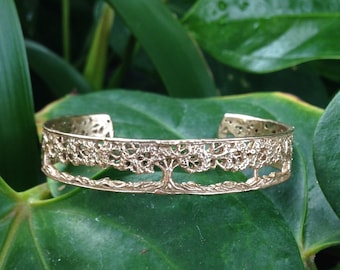 May include: A gold-toned metal cuff bracelet featuring an openwork design of a tree with a textured canopy and a solid base band. The bracelet has a polished finish and a wide, circular opening, showcasing intricate detailing across the metal surface.