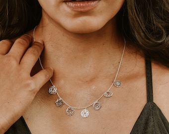 May include: A silver chain necklace featuring seven small, circular charms, each engraved with a distinct geometric chakra symbol. The necklace includes a matching silver earring with a circular, intricate, openwork geometric design.