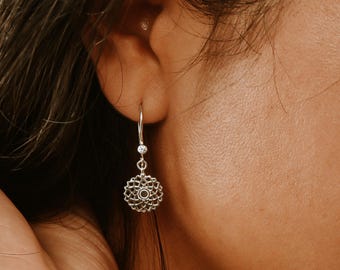 May include: A silver drop earring featuring a circular, openwork mandala-style pendant with a small, clear, round-cut stone set at the top of the hook and a matching stone at the center of the intricate, geometric, floral-patterned metal design.