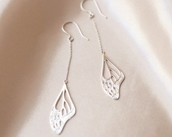 May include: A pair of silver-colored dangle earrings. Each earring features a delicate, openwork design resembling a butterfly wing. The wings are suspended from a chain and a curved ear wire. The earrings are displayed on a satin-like, beige background.