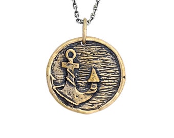 May include: A round, bronze-colored pendant necklace featuring an anchor design. The anchor is set against a textured background, and the pendant hangs from a silver-colored chain. The nautical-themed jewelry piece is displayed against a white background.