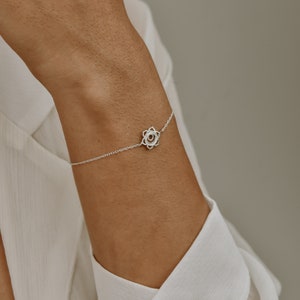 Puede incluir: Una delicada pulsera de plata con un dije floral. El dije tiene un diseño calado con un pequeño centro circular. La pulsera se lleva en la muñeca y la cadena es fina y elegante. La pulsera es una joya.