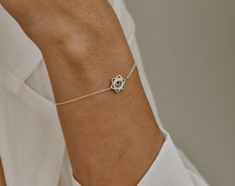 May include: A delicate silver bracelet featuring a floral charm. The charm has an openwork design with a small, circular center. The bracelet is worn on a wrist, and the chain is fine and elegant. The bracelet is a piece of jewelry.
