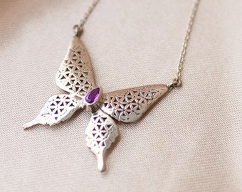May include: A silver butterfly necklace with a purple gemstone in the center. The butterfly is intricately designed with a geometric pattern.