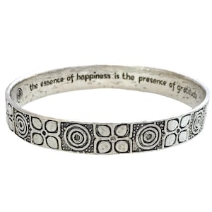 Könnte beinhalten: Silbernes Armreif-Armband mit einem sich wiederholenden Muster aus Kreisen und Quadraten. Die Inschrift auf dem Armband lautet "the essence of happiness is the presence of gratitude".