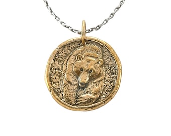 May include: A gold-toned pendant necklace featuring a detailed bear head design. The pendant is round and has a textured surface. The chain is silver-toned.