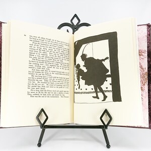 May include: An open book with a decorative cover, displayed on a black metal stand. The book's pages feature text and a black silhouette illustration of a person carrying another person. The cover has a patterned design.