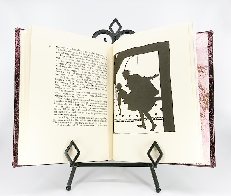 May include: An open book with a decorative cover, displayed on a black metal stand. The book's pages feature text and a black silhouette illustration of a person carrying another person. The cover has a patterned design.
