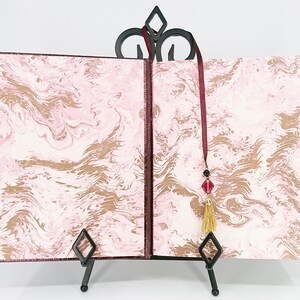 May include: An open book with pink and gold marbled pages, displayed on a black metal stand. The book has a burgundy cover and a burgundy ribbon with a tassel and bead embellishments. The stand has a decorative top.