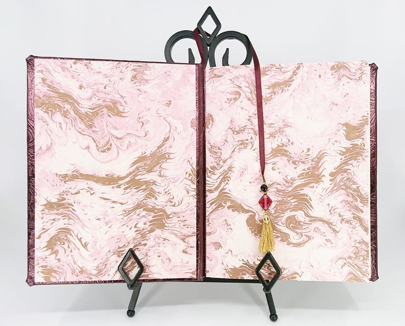 May include: An open book with pink and gold marbled pages, displayed on a black metal stand. The book has a burgundy cover and a burgundy ribbon with a tassel and bead embellishments. The stand has a decorative top.