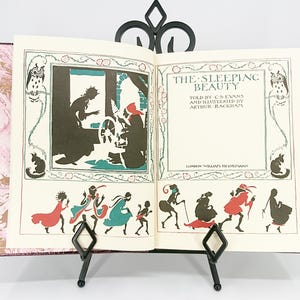 May include: An open book titled "The Sleeping Beauty" with illustrations by Arthur Rackham. The book is propped up on a black metal stand. The pages feature black and colored illustrations of characters and scenes from the fairy tale.