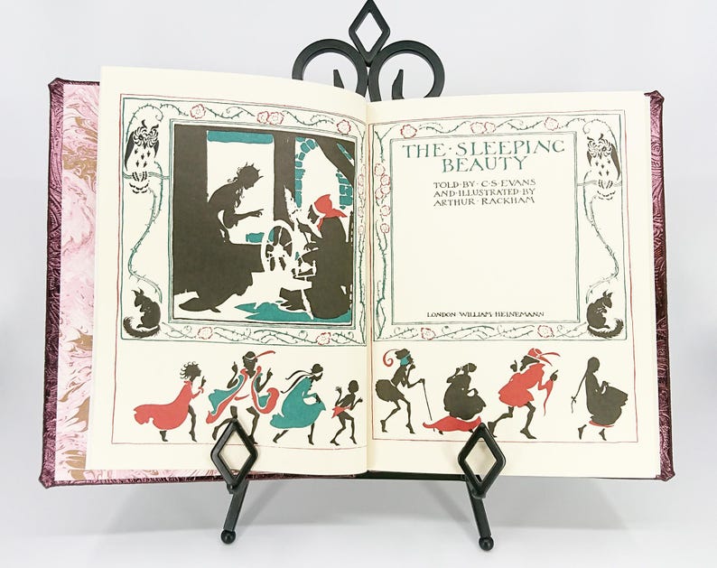 May include: An open book titled "The Sleeping Beauty" with illustrations by Arthur Rackham. The book is propped up on a black metal stand. The pages feature black and colored illustrations of characters and scenes from the fairy tale.