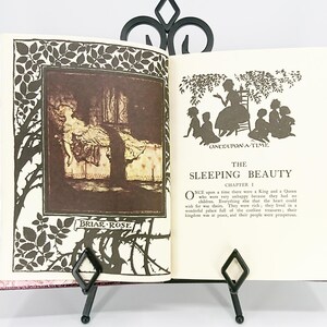 May include: An open book with illustrations from "The Sleeping Beauty" fairy tale, held by a black metal stand. The book's pages feature text and sepia-toned images, including a silhouette of a woman and children. The cover is a marbled pink.