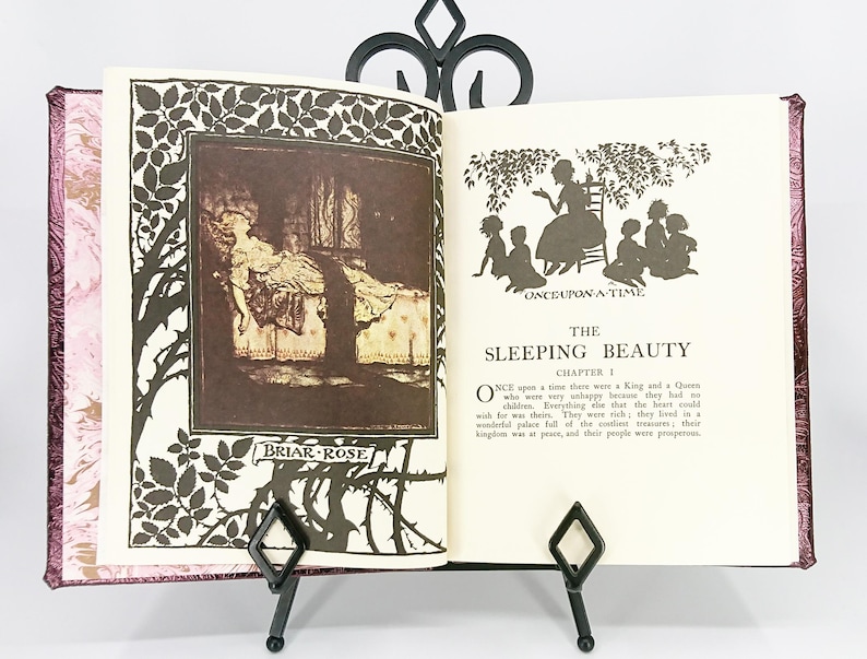 May include: An open book with illustrations from "The Sleeping Beauty" fairy tale, held by a black metal stand. The book's pages feature text and sepia-toned images, including a silhouette of a woman and children. The cover is a marbled pink.
